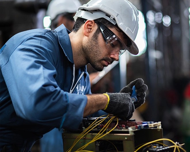 Person in a white safety helmet and blue uniform using a screwdriver on an electronic device with multiple wires, in an industrial setting.