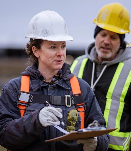Four construction workers in safety gear standing outdoors, with one holding a clipboard and speaking to the group.