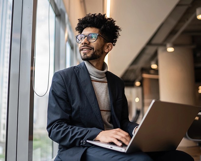 Individual in a dark blazer and striped turtleneck sitting near a large window with a city view, typing on a laptop in a modern indoor space.