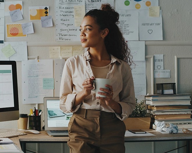 Individual in a light shirt and brown pants holding a white mug, standing near a desk with a laptop, books, and a wall covered in notes, charts, and a whiteboard.