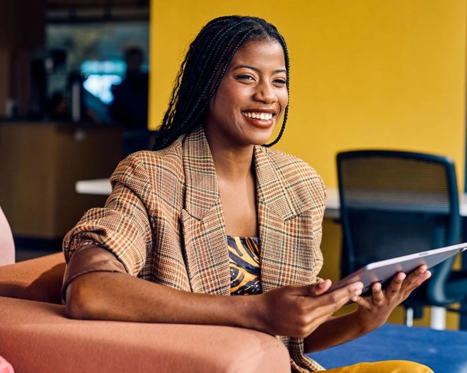 Individual with long braided hair wearing a plaid blazer, smiling while holding a tablet on a peach-coloured couch in a casual workspace with a yellow wall.
