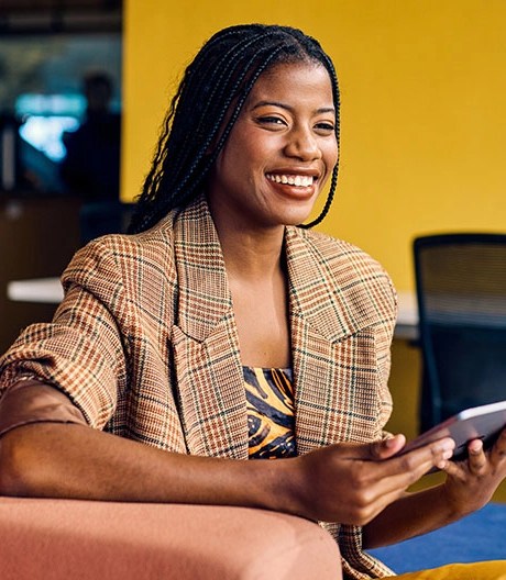 Individual with long braided hair wearing a plaid blazer, smiling while holding a tablet on a peach-coloured couch in a casual workspace with a yellow wall.