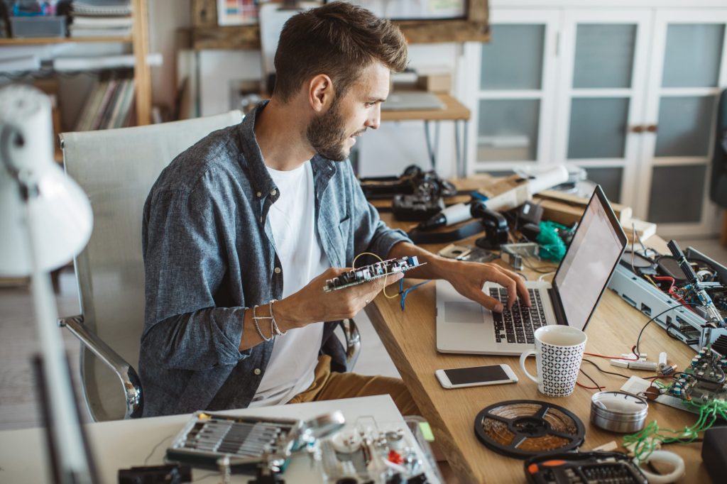 Individual seated at a desk holding a circuit board and using a laptop, surrounded by electronic components, tools, a coffee mug, and a notebook.