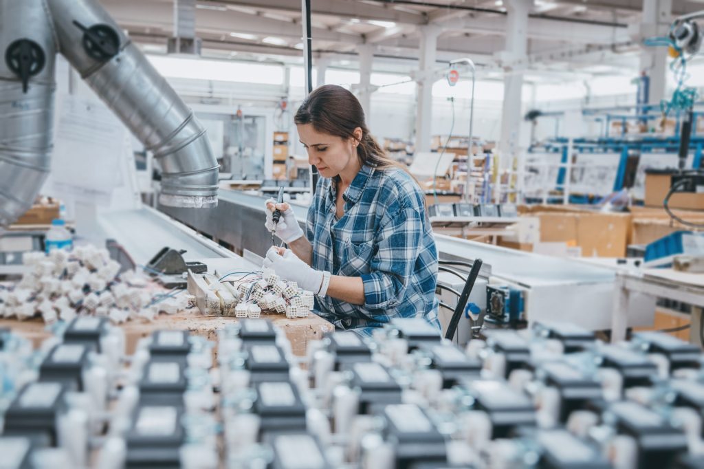 Person in a blue plaid shirt and white gloves assembling electronic components at a workstation, with rows of devices and a spacious factory in the background.