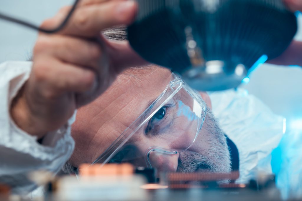 Individual in safety goggles and a white protective suit handling a large lens above a circuit board in a laboratory or technical environment.