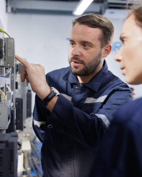 Two people in dark blue uniforms with reflective stripes standing in front of an electrical control panel, with one pointing at a component while the other observes.