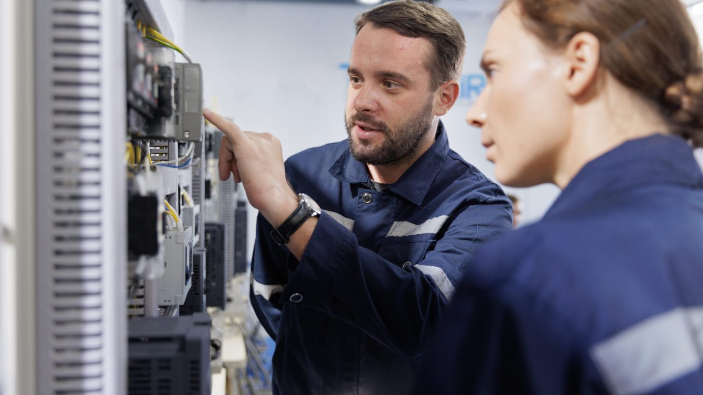 Two people in dark blue uniforms with reflective stripes standing in front of an electrical control panel, with one pointing at a component while the other observes.