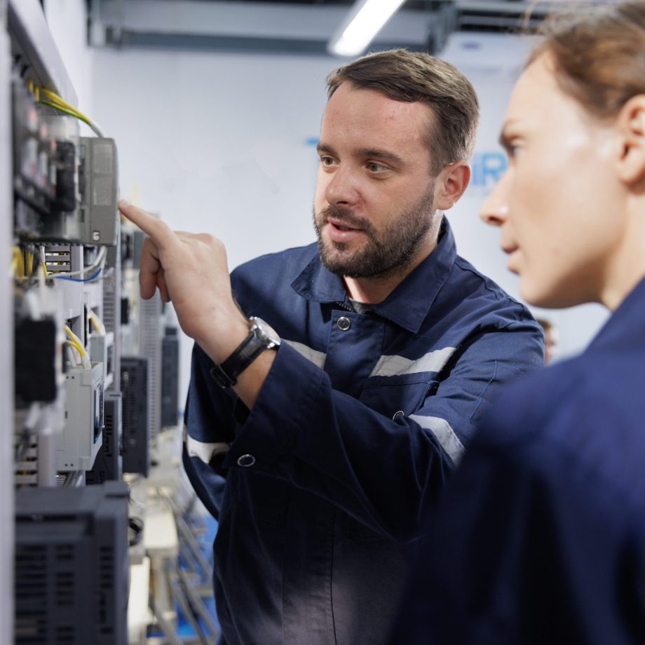 Two people in dark blue uniforms with reflective stripes standing in front of an electrical control panel, with one pointing at a component while the other observes.
