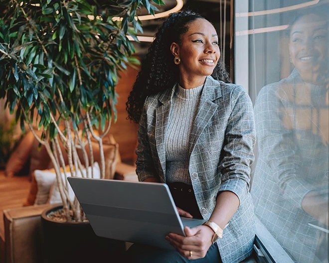 Individual in a grey plaid blazer and turtleneck sweater holding a laptop and looking out a large window, seated in a modern indoor space with a potted plant.