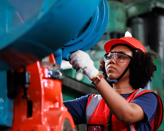Person in a red hard hat and reflective vest inspecting a blue component on an industrial machine with an orange base, in a factory setting.