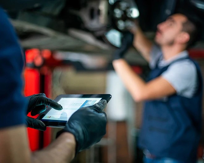 Individual wearing black gloves holding a tablet displaying text and graphics, while another person works on the underside of a vehicle in a workshop.