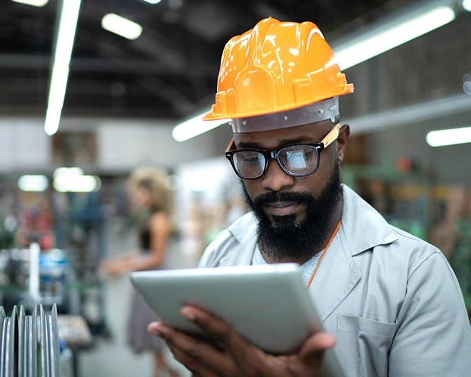 Person in an orange safety helmet and gray uniform looking at a tablet in an industrial environment with blurred machinery and another worker in the background.