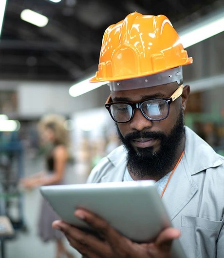 Person in an orange safety helmet and gray uniform looking at a tablet in an industrial environment with blurred machinery and another worker in the background.