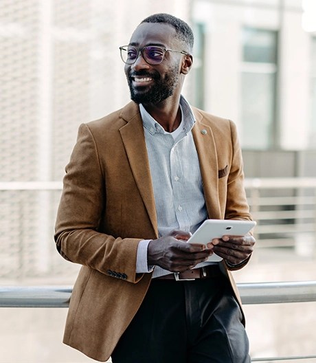Individual in a brown blazer and light blue shirt standing near a metal railing in an urban setting, holding a tablet with modern architecture in the background.