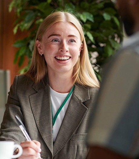 Woman in a business suit with a lanyard smiling and holding a pen during a conversation, seated at a table with a white coffee cup and a green plant in the background.