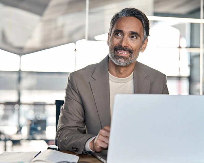Man in a light brown blazer seated at a desk with an open laptop and notebook, looking to the side in a modern office with large windows.