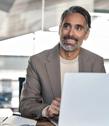 Man in a light brown blazer seated at a desk with an open laptop and notebook, looking to the side in a modern office with large windows.