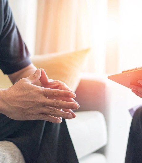 Two individuals in a counseling or consultation setting, with one person seated on a couch clasping their hands and the other holding a clipboard, taking notes.