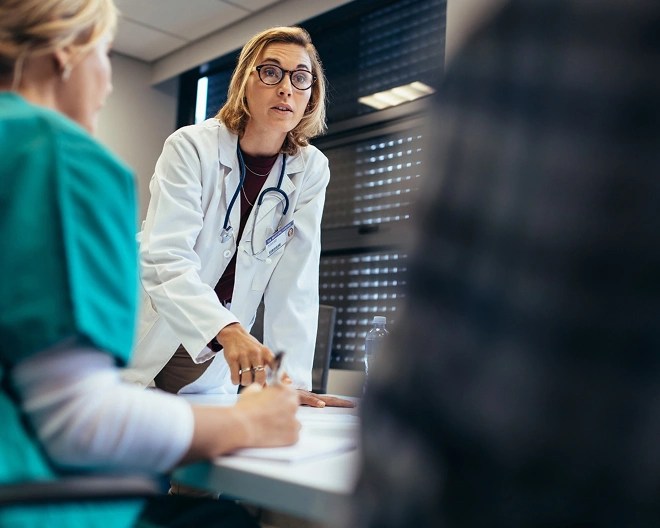 Group of healthcare professionals in a meeting, with one person in a white lab coat speaking and gesturing, and another in green scrubs writing on a notepad.