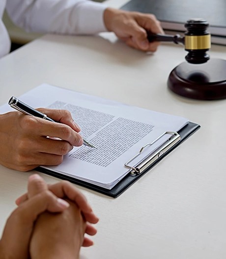 Person in formal attire writing on a clipboard while another individual sits with clasped hands, with a gavel and closed book on the table in a legal setting.