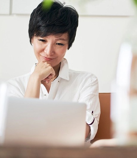 Individual in a white shirt sitting on a couch, resting their chin on their hand while looking at a laptop, with blurred objects in the foreground and a minimalistic background.