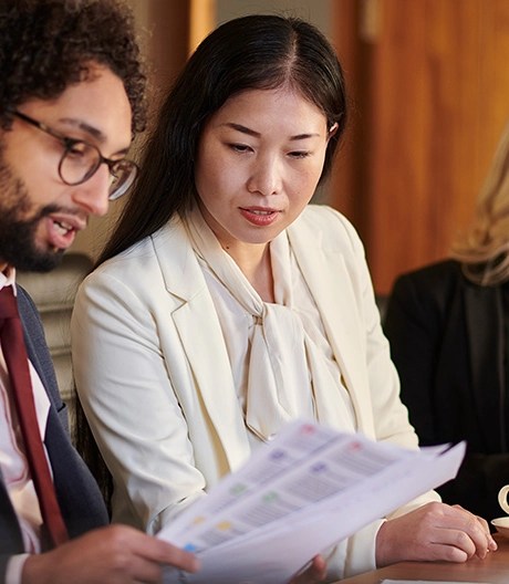 Three individuals seated at a table in a professional setting, with one person reviewing a document containing colorful charts, another observing attentively, and a third interacting with a smartphone.