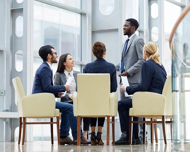 Five professionally dressed individuals in a modern office, with one person standing and presenting to four others seated in cream-colored chairs, reviewing documents.