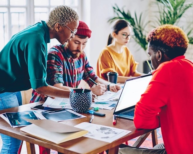Four people gathered around a wooden table with documents, charts, a laptop, and a coffee cup, in a bright room with large windows and indoor plants.
