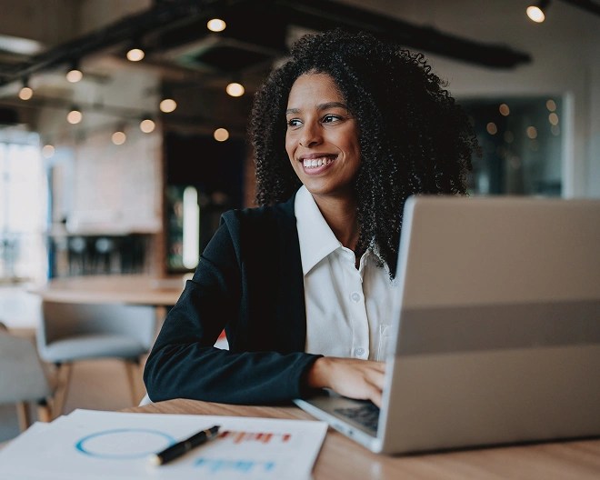 Individual seated at a table in a modern office, smiling and looking to the side with an open laptop and documents displaying charts and graphs.
