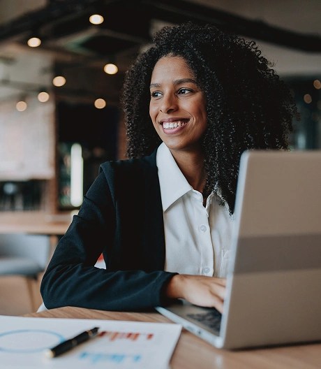Individual seated at a table in a modern office, smiling and looking to the side with an open laptop and documents displaying charts and graphs.