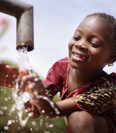 Young child smiling while washing hands under a running water tap, wearing a patterned dress and gold earrings with greenery in the background.