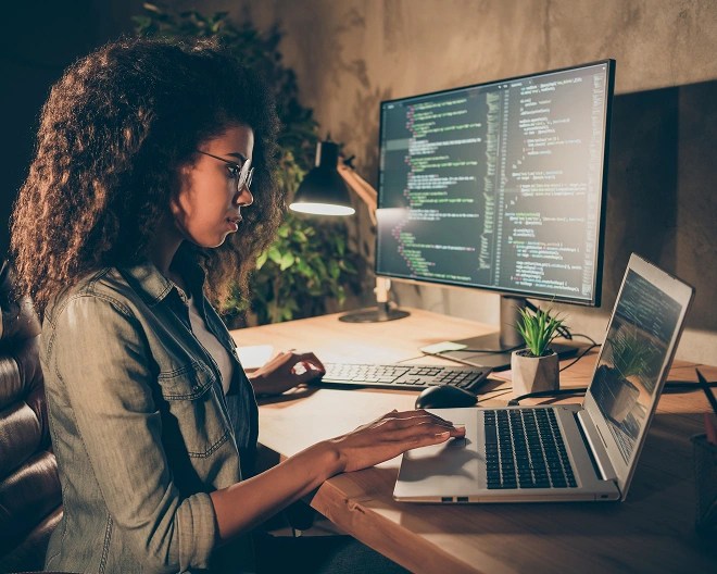Individual working at a desk with a laptop and external monitor displaying lines of code.