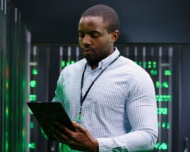Individual in a checkered shirt and ID badge holding a tablet in a server room with green LED-lit racks.