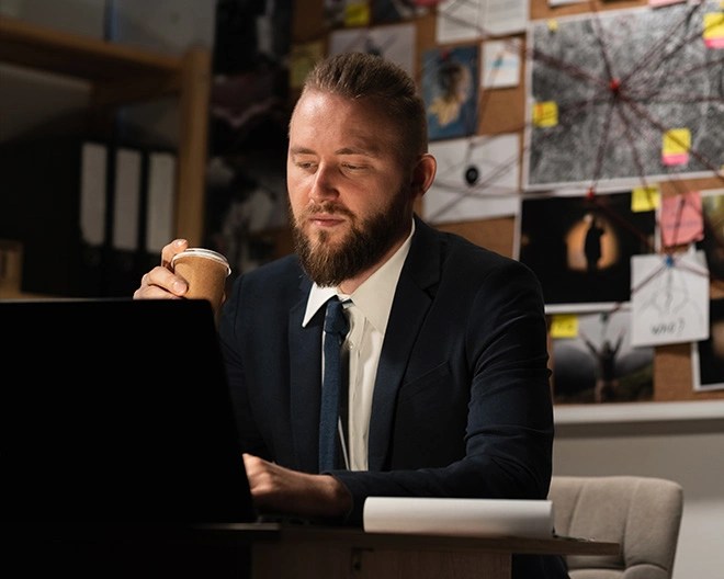 Man in a suit holding a paper coffee cup and looking at a laptop, seated in front of a bulletin board filled with photos, notes, and string connections.