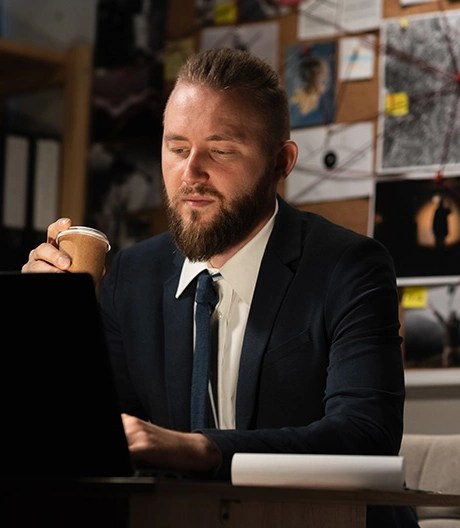 Man in a suit holding a paper coffee cup and looking at a laptop, seated in front of a bulletin board filled with photos, notes, and string connections.
