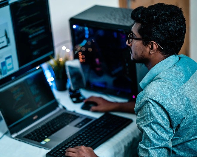 Person working at a desk with a laptop, external keyboard, mouse, and large monitor displaying code and a design interface, next to a desktop tower with RGB lighting.