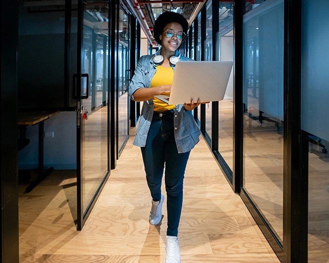 Individual wearing a yellow top, denim shirt, and headphones around the neck walking through a glass-walled hallway while holding an open laptop.