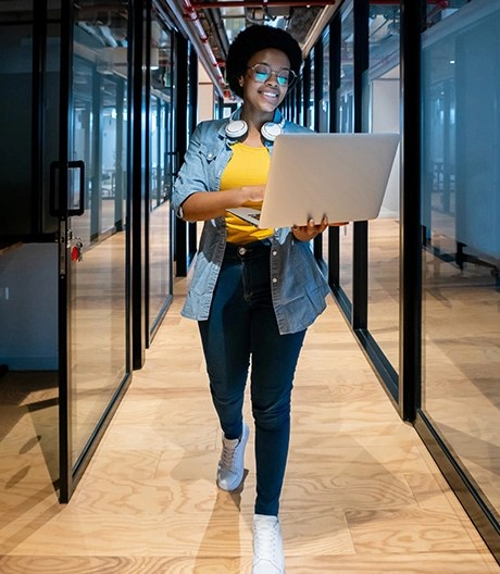 Individual wearing a yellow top, denim shirt, and headphones around the neck walking through a glass-walled hallway while holding an open laptop.