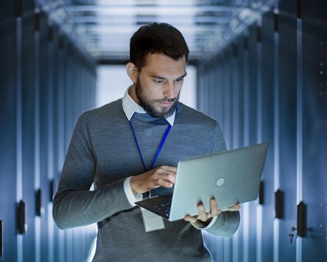 Individual in a gray sweater and ID badge using a laptop in a server room with rows of blue-lit server racks.