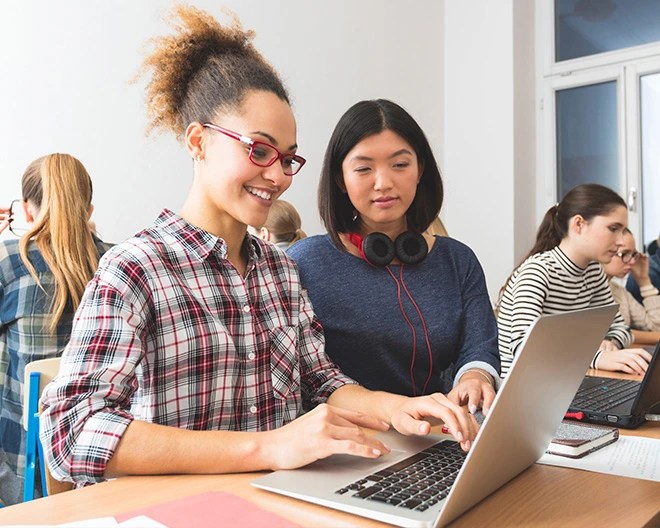 Two students working together on a laptop in a classroom, with others in the background also using laptops or talking, in a bright room with large windows.