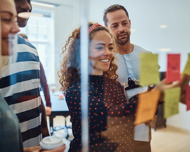 Four people standing in front of a glass wall covered in colourful sticky notes, with one person pointing and others observing or holding a clipboard and coffee cup.