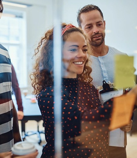 Four people standing in front of a glass wall covered in colourful sticky notes, with one person pointing and others observing or holding a clipboard and coffee cup.