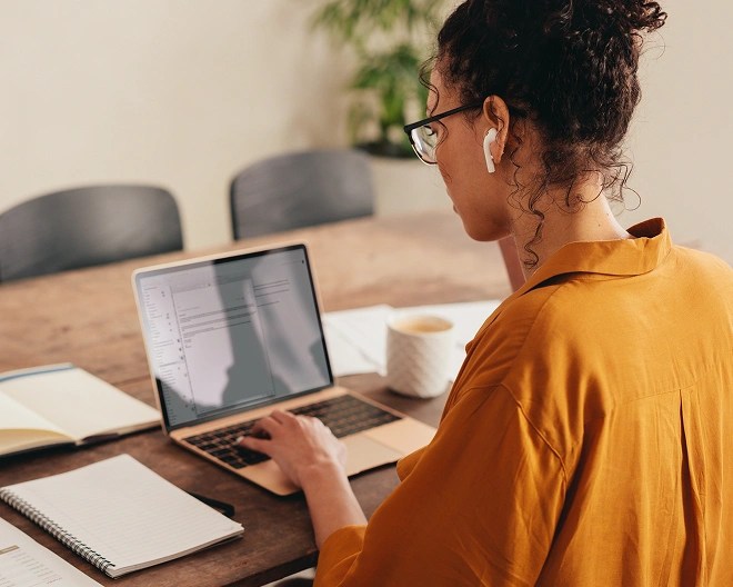 Individual in a mustard-colored shirt using a laptop at a wooden table with notebooks, papers, a ceramic mug, and earbuds in, in a casual indoor setting.