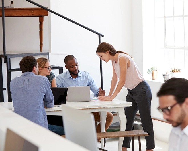 Five people in a modern office seated around a white table with laptops, engaged in a collaborative discussion, with one person standing and leaning over the table.