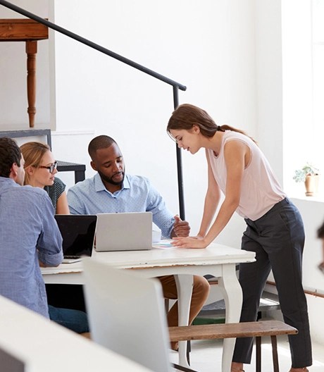 Five people in a modern office seated around a white table with laptops, engaged in a collaborative discussion, with one person standing and leaning over the table.