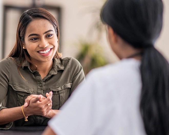 Two individuals talking indoors, with one person in a green button-up shirt smiling and facing the camera, and the other wearing a white top with their back to the camera.