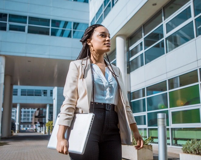 Individual in business attire holding a silver laptop while walking through an urban area with modern glass architecture.