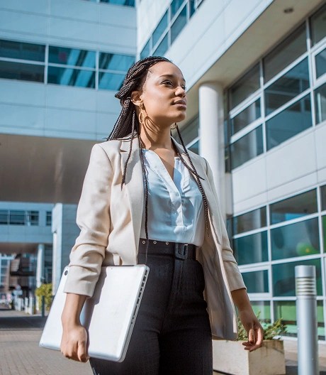 Individual in business attire holding a silver laptop while walking through an urban area with modern glass architecture.