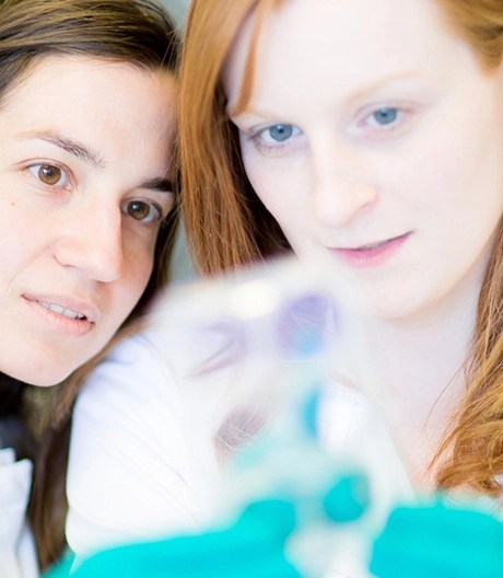 Two people in white lab coats closely examining a transparent object held by one of them wearing green gloves, in a laboratory setting.