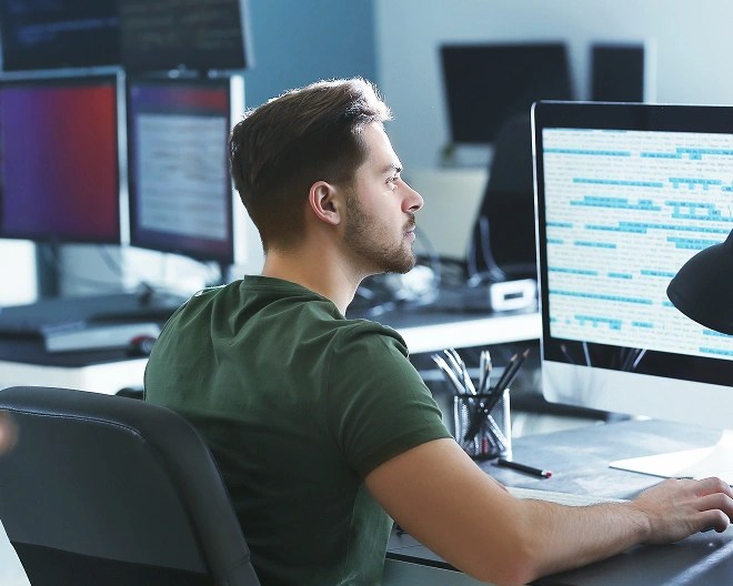 Individual seated at a desk in front of multiple monitors displaying code or data, in a clean and organised tech workspace.